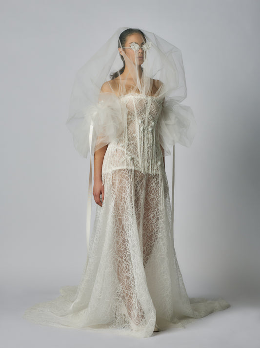 Model wearing a white lace wedding dress with a sheer overlay against a plain background