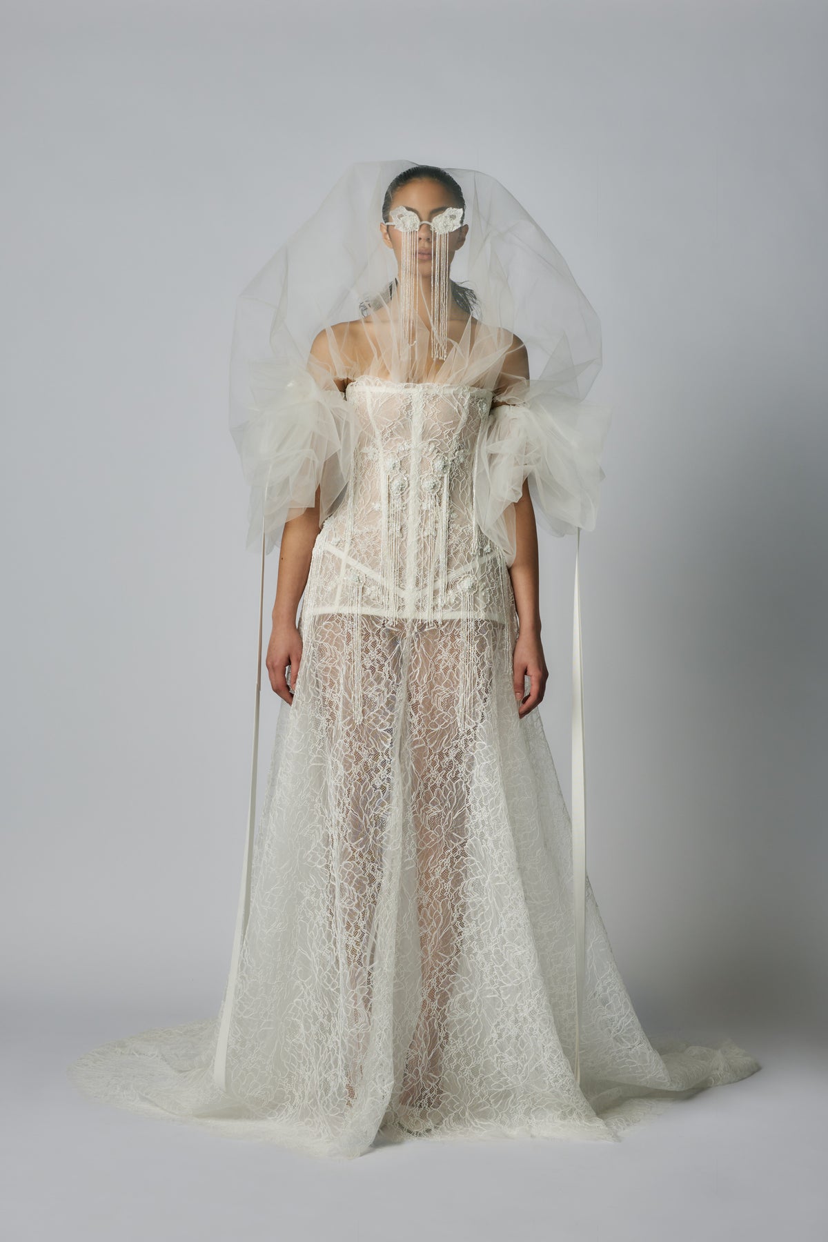 Model wearing a white lace wedding dress with a sheer veil against a plain background